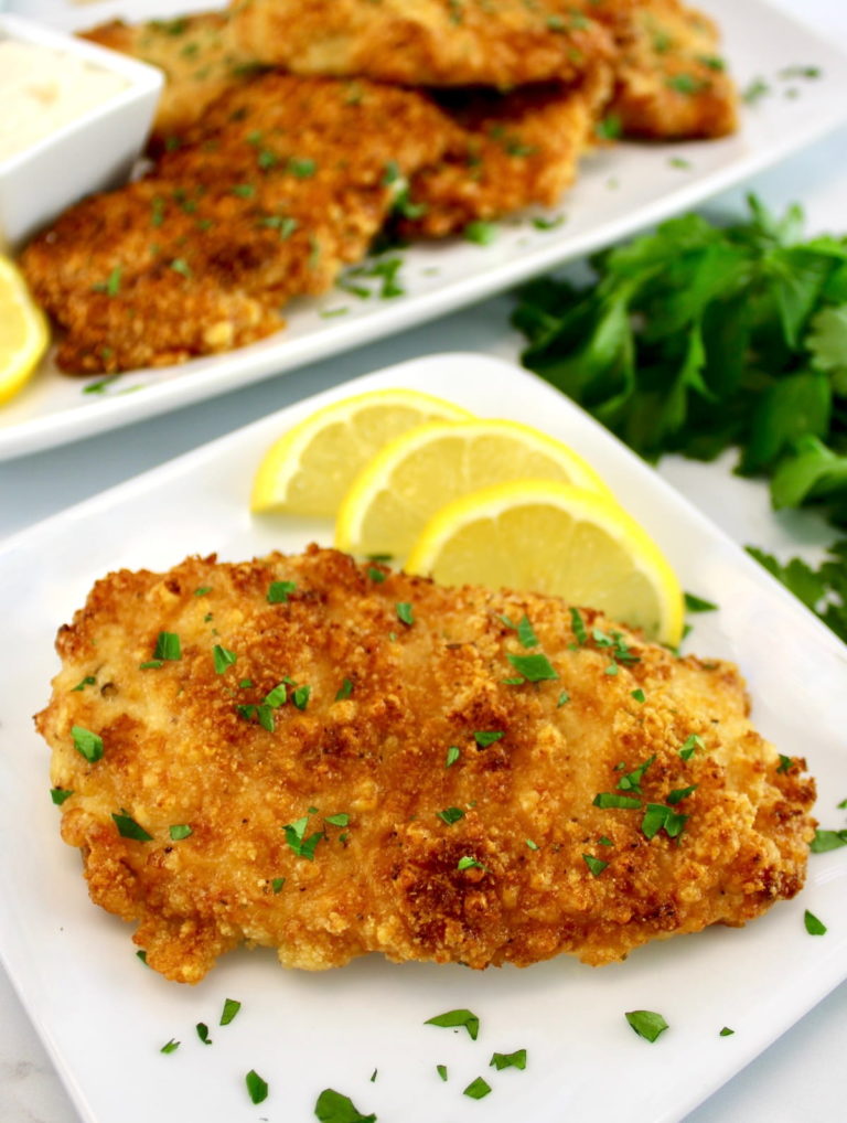 closeup of piece of Air Fryer Parmesan Crusted Chicken on white plate with lemon slices and more chicken in back