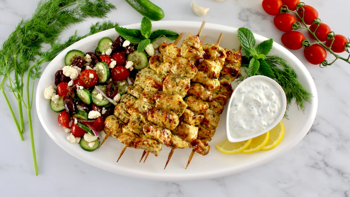 Chicken Souvlaki skewers with greek salad with dipping sauce on a white plate