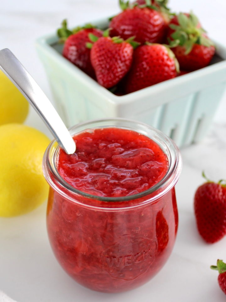 Keto Strawberry Sauce in open glass jar with spoon and strawberries in back