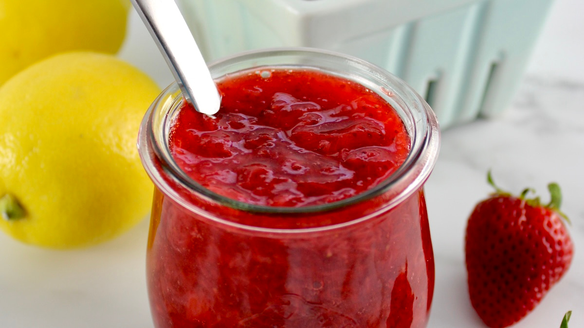closeup of Keto Strawberry Sauce in open glass jar with spoon and lemons in back