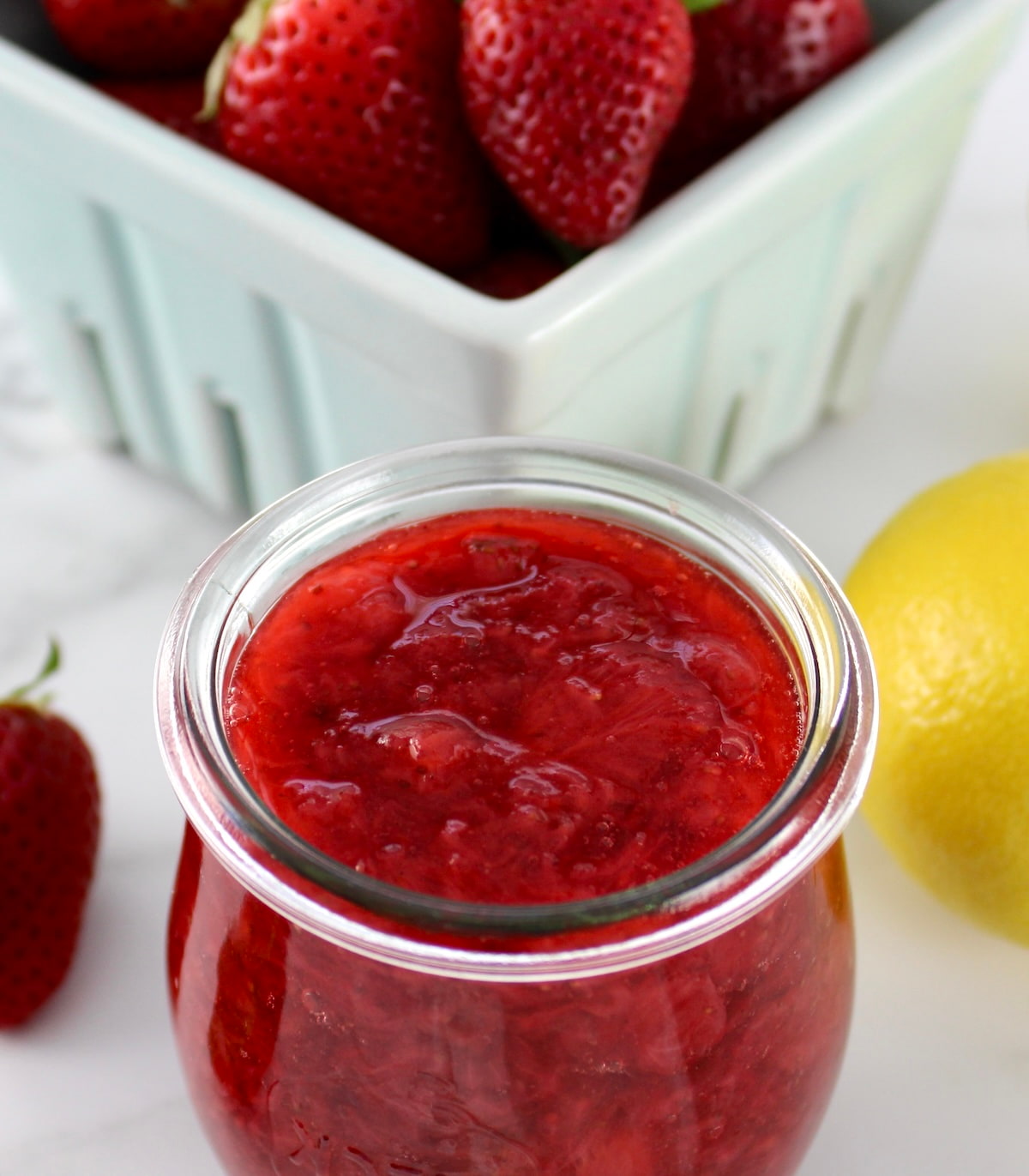 closeup of Keto Strawberry Sauce in open glass jar with fresh strawberries and lemons in back