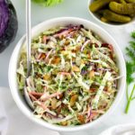 Dill Pickle Coleslaw in white bowl with silver serving spoon and cabbage in back