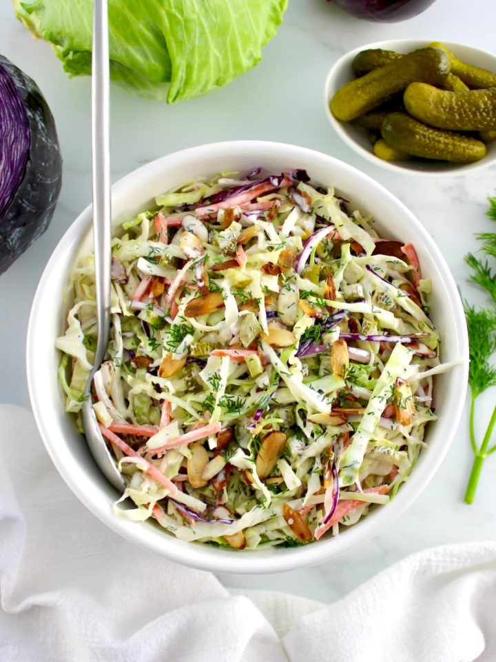 Dill Pickle Coleslaw in white bowl with silver serving spoon and cabbage in back