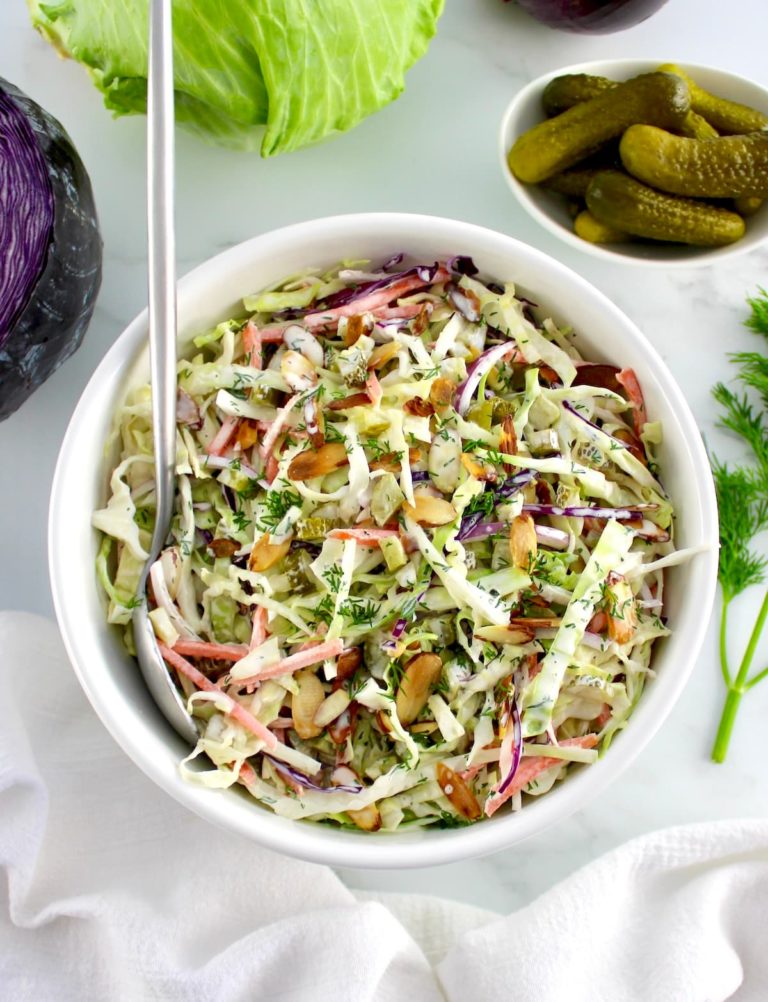 Dill Pickle Coleslaw in white bowl with silver serving spoon and cabbage in back