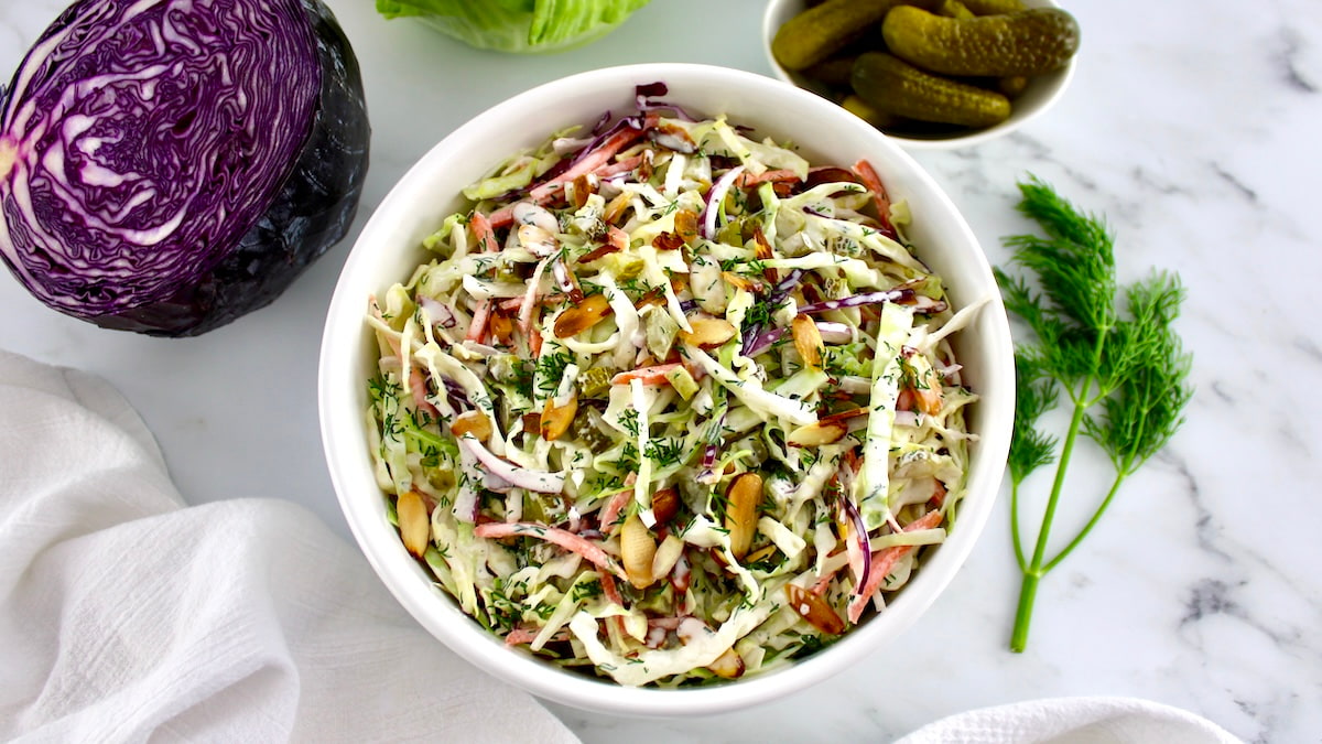 overhead view of Dill Pickle Coleslaw in white bowl with cabbage and pickles in background