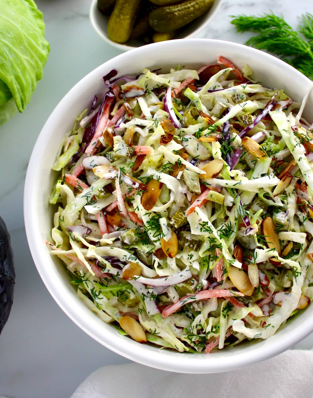Dill Pickle Coleslaw in white bowl with cabbage and pickles in back