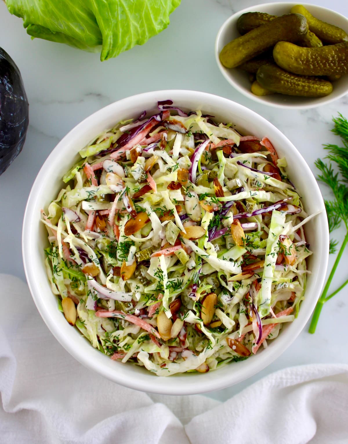overhead view of Dill Pickle Coleslaw in white bowl with cabbage and pickles in background