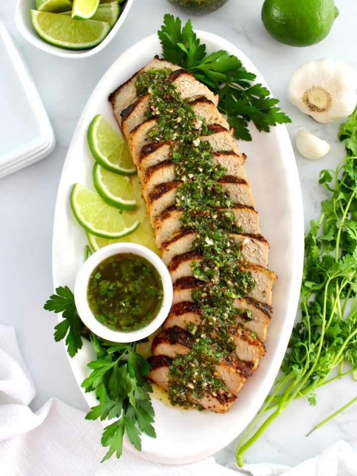 Grilled Pork Tenderloin Chimichurri sliced on white platter with sauce in white cup and lime slices