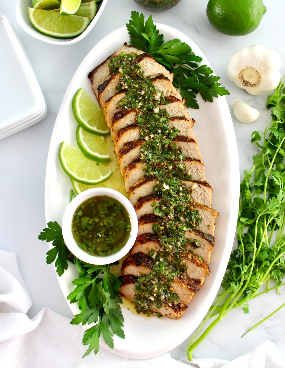 Grilled Pork Tenderloin Chimichurri sliced on white platter with sauce in white cup and lime slices