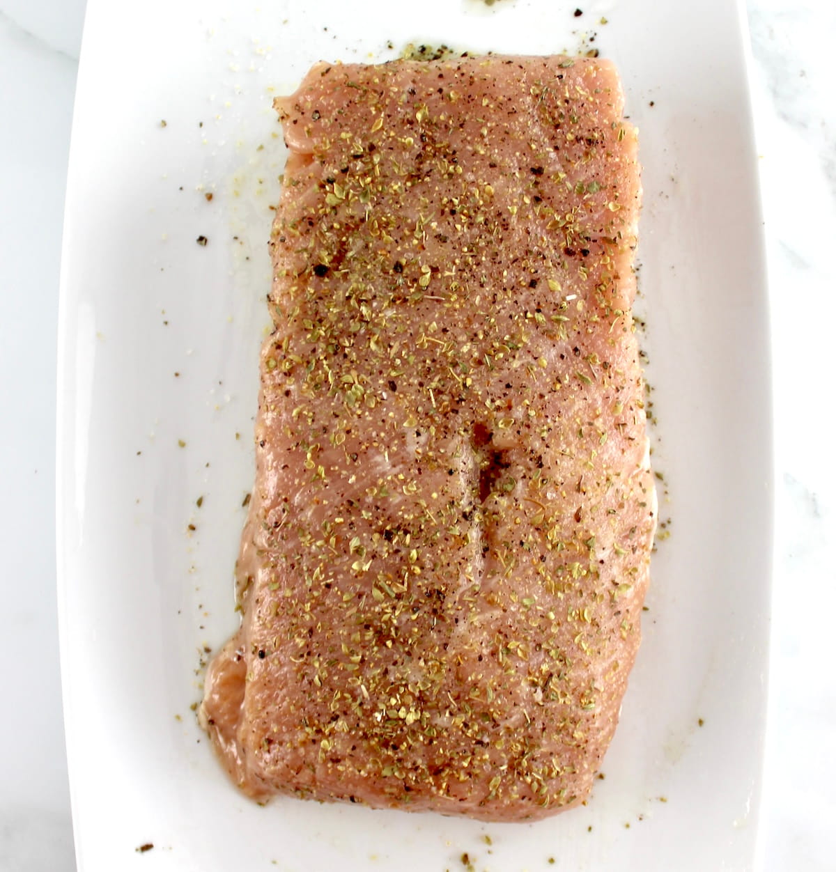 raw pork tenderloin on white plate with olive oil, dried oregano, salt and pepper