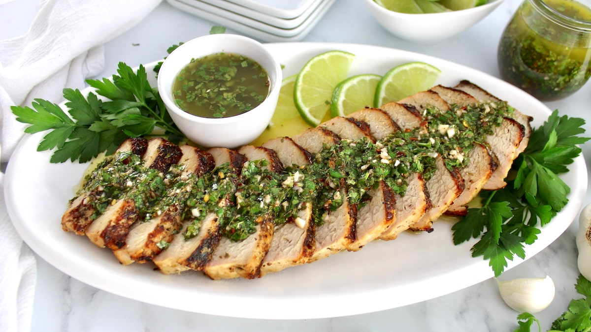Grilled Pork Tenderloin with Chimichurri sliced on white plate with sauce in cup and lime slices