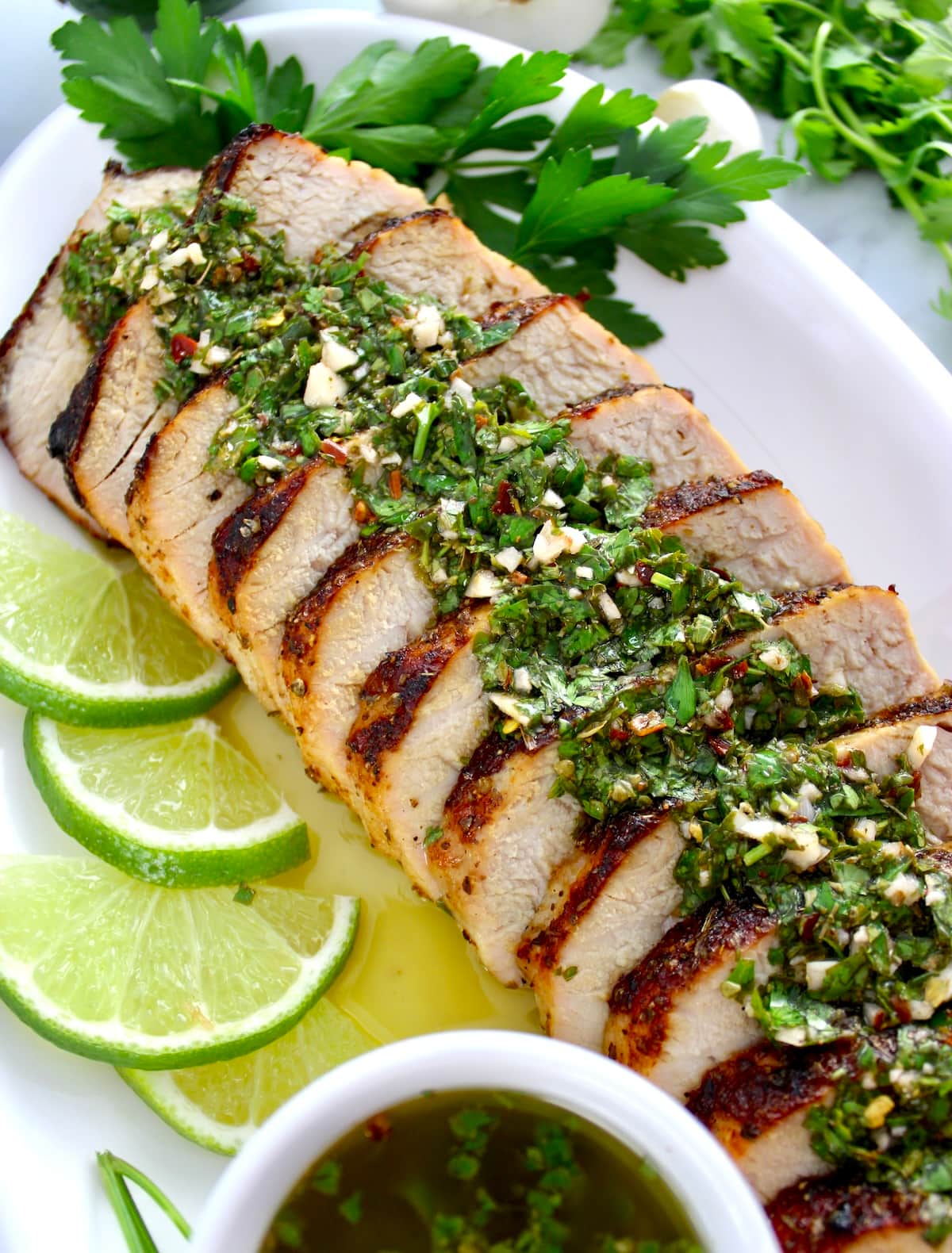 Grilled Pork Tenderloin with Chimichurri sauce on top sliced on white plate and lime slices
