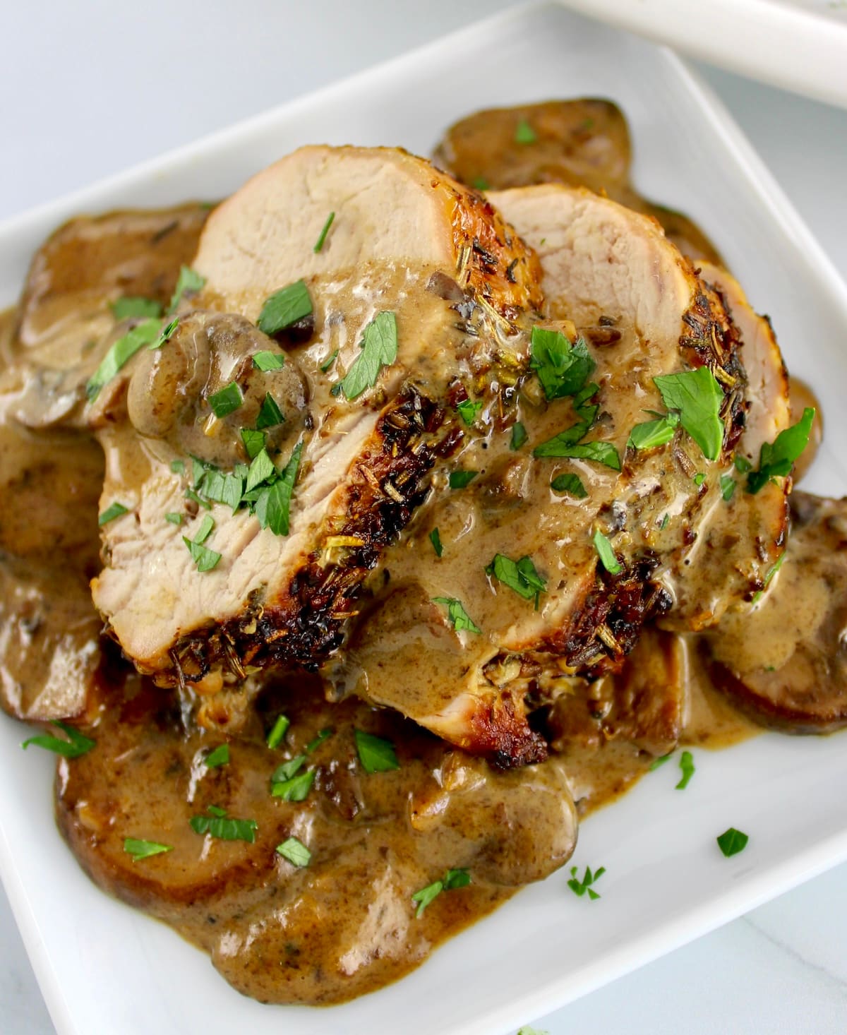 closeup of Herb Crusted Pork Tenderloin with Mushroom Gravy on white plate with chopped parsley