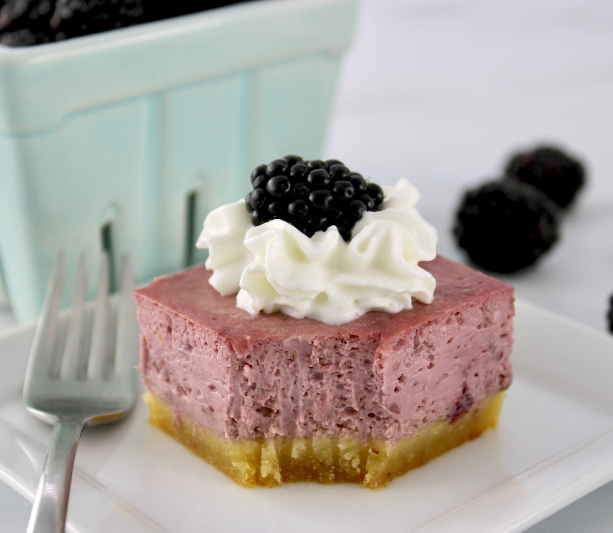 Blackberry Cheesecake Bar on white plate with whip cream and blackberry on top with bite missing and fork on side