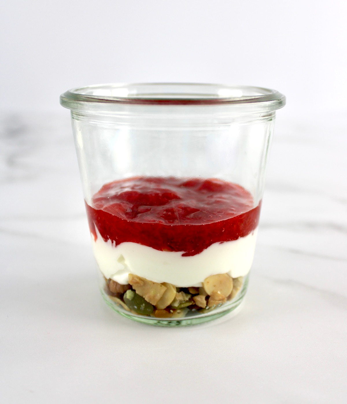 side view of open glass jar with keto granola, Greek yogurt and strawberry sauce in layers