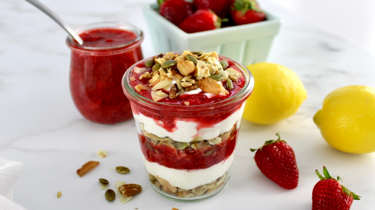 sideview of Keto Strawberry Granola Parfait in open glass jar with strawberries and lemon in back