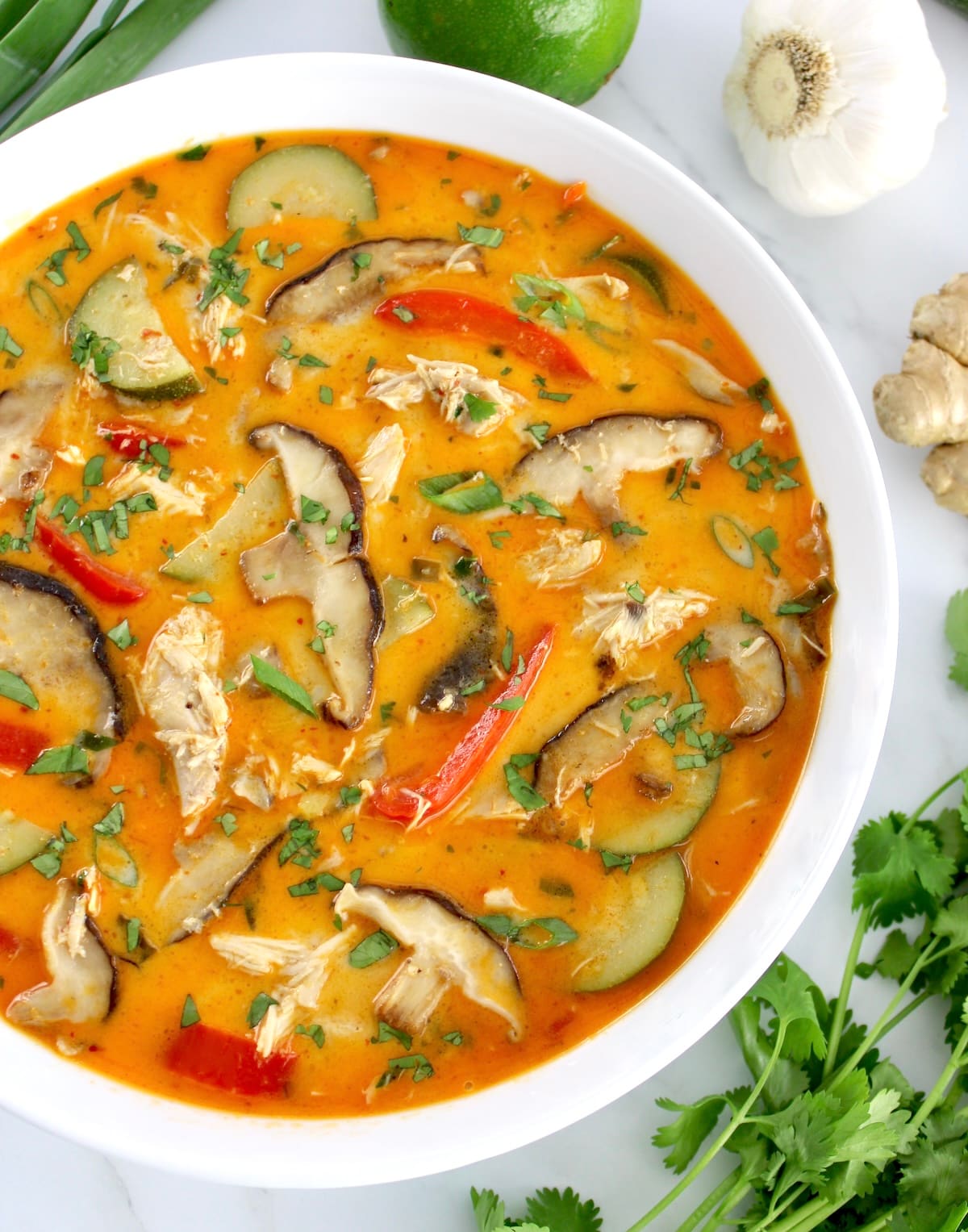 overhead view of Thai Coconut Curry Chicken Soup in white bowl with fresh cilantro and veggies around