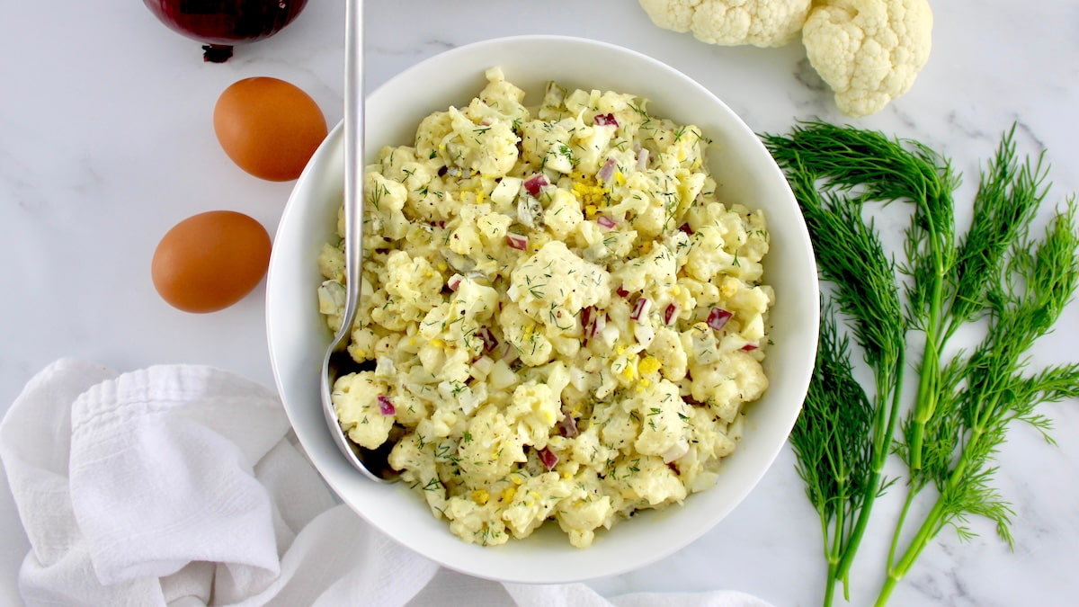 Cauliflower Potato Salad in white bowl with serving spoon and dill, cauliflower and eggs in background