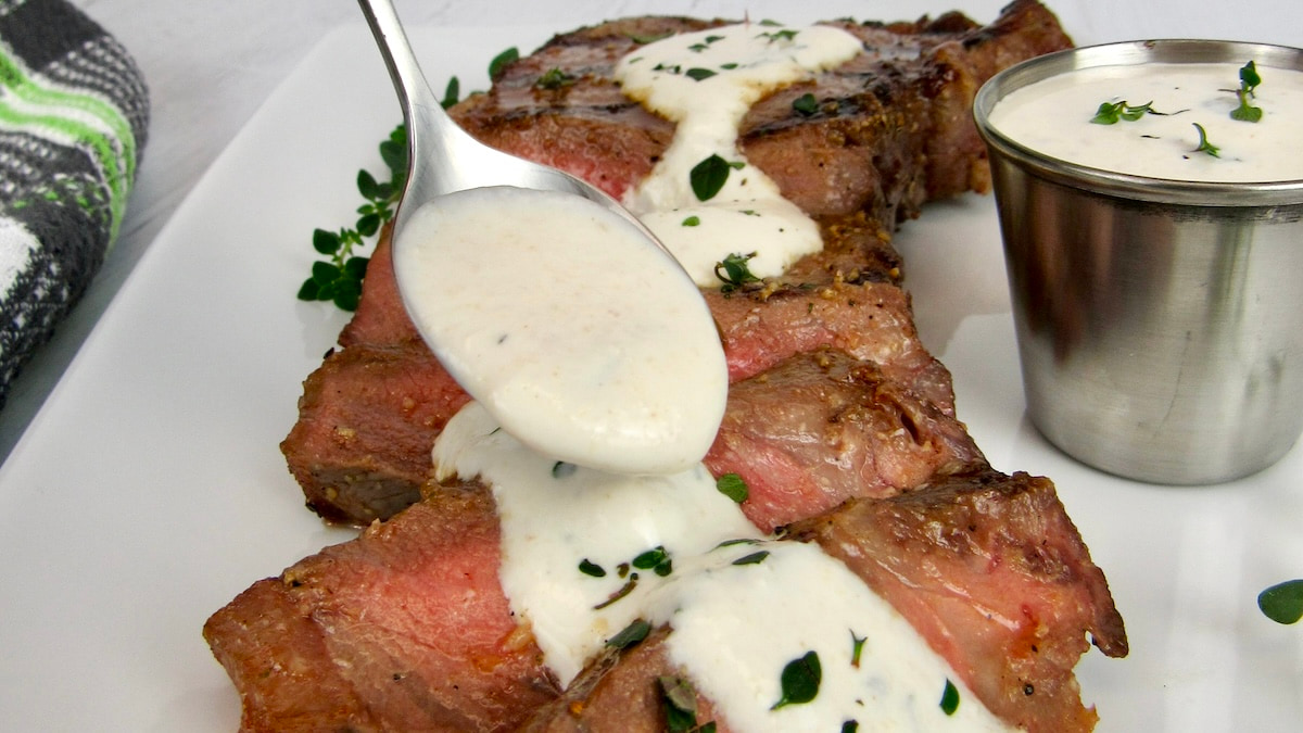 Creamy Horseradish Sauce being spooned over sliced steak on white plate