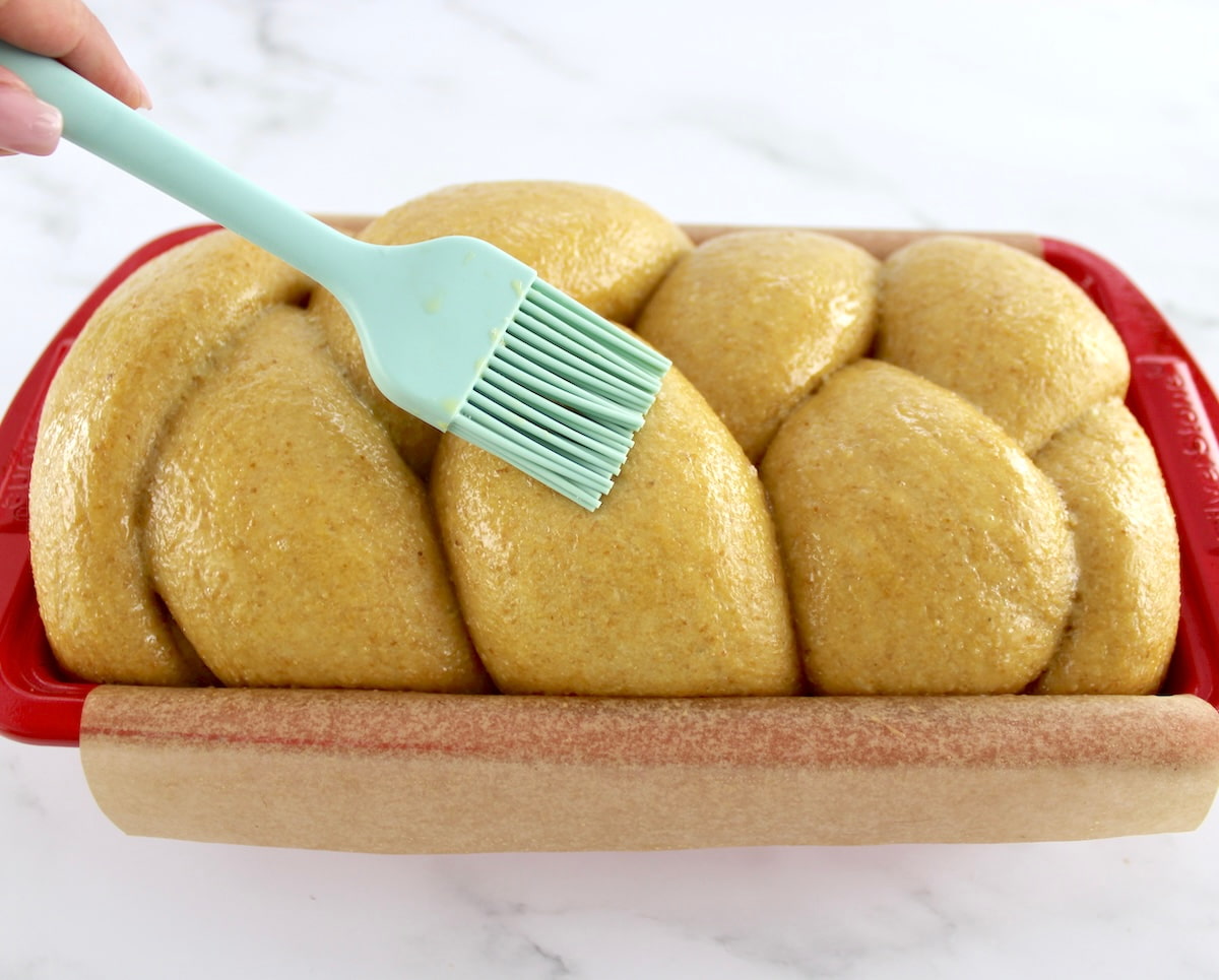Keto Brioche Bread braid dough in red loaf pan lined with parchment and being brushed with egg wash