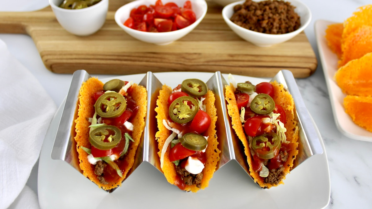 3 Keto Cheese Taco Shells in metal taco holder with toppings in white bowls in back