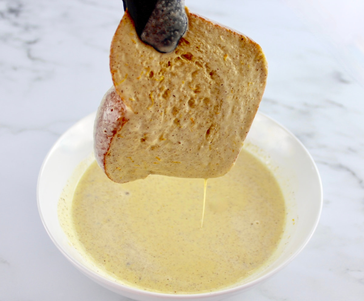slice of french toast being dipped in egg mixture held up by tongs