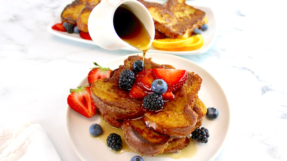 Keto French Toast on white plate with fresh berries on top and maple syrup being poured over
