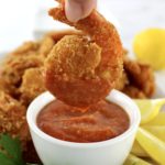 dipping fried shrimp in cocktail sauce in white bowl with lemon and parsley on side