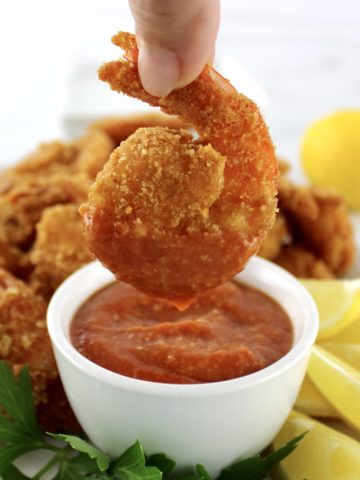 dipping fried shrimp in cocktail sauce in white bowl with lemon and parsley on side