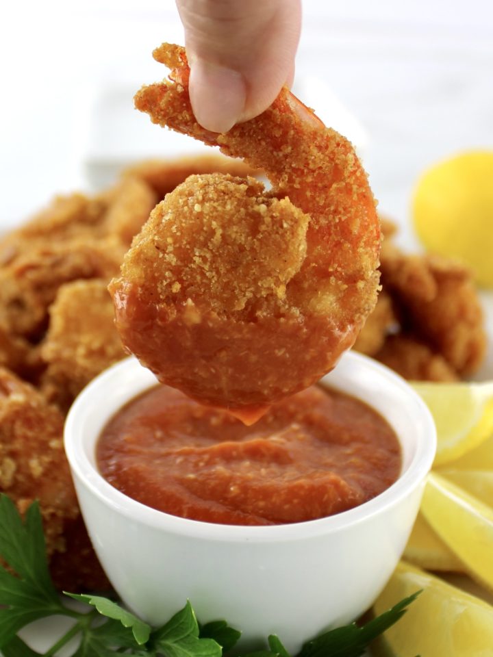 dipping fried shrimp in cocktail sauce in white bowl with lemon and parsley on side