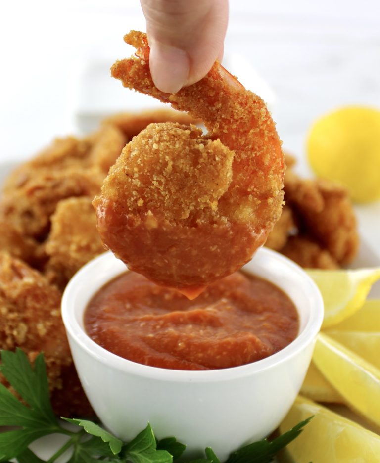dipping fried shrimp in cocktail sauce in white bowl with lemon and parsley on side