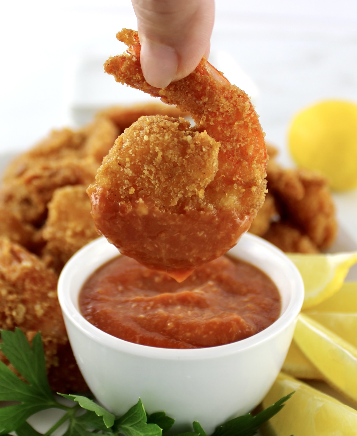 dipping fried shrimp in cocktail sauce in white bowl with lemon and parsley on side