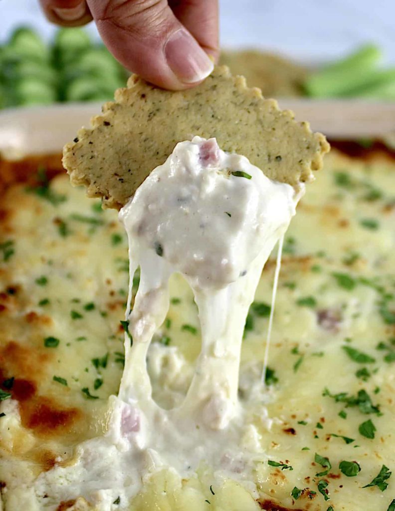 Chicken Cordon Bleu Dip being scooped out with cracker and cheese pulling