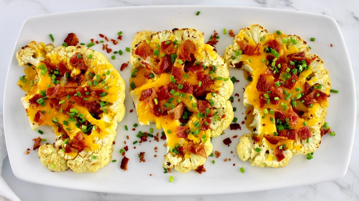 3 Loaded Cauliflower Steaks on white platter with crumbled bacon and chives on top