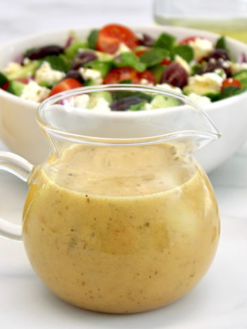 Greek Salad Dressing in glass pitcher with salad in back