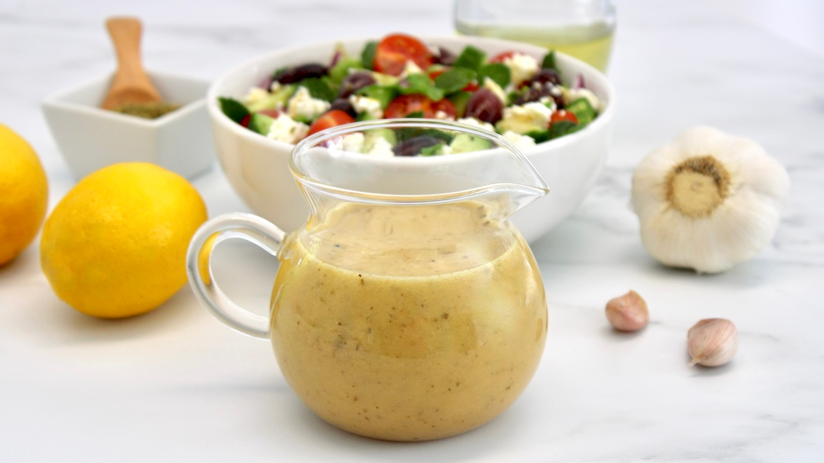 Greek Salad Dressing in glass pitcher with salad in back