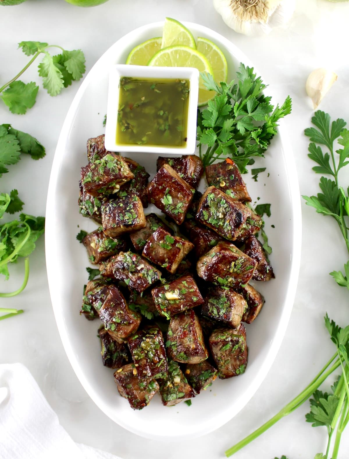 overhead view of Chimichurri Steak Bites on white oval platter with sauce on side with lime slices
