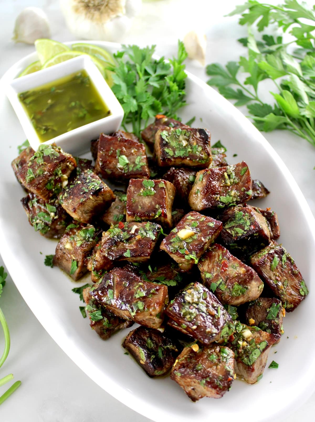 Chimichurri Steak Bites on white oval platter with sauce and limes on side