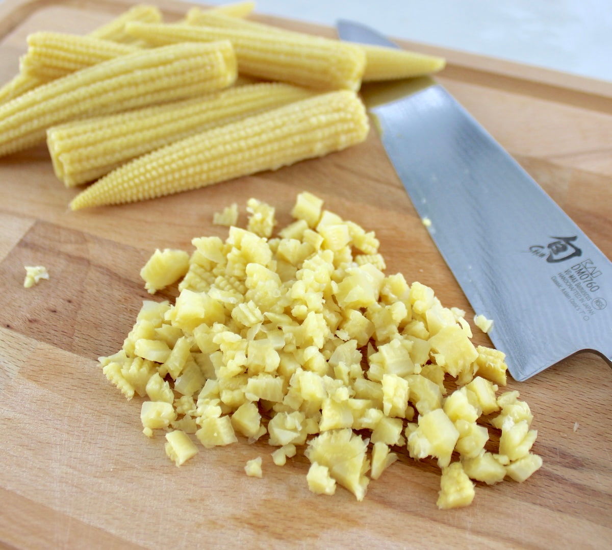 chopped baby corn on cutting board with knife and whole baby corn in back