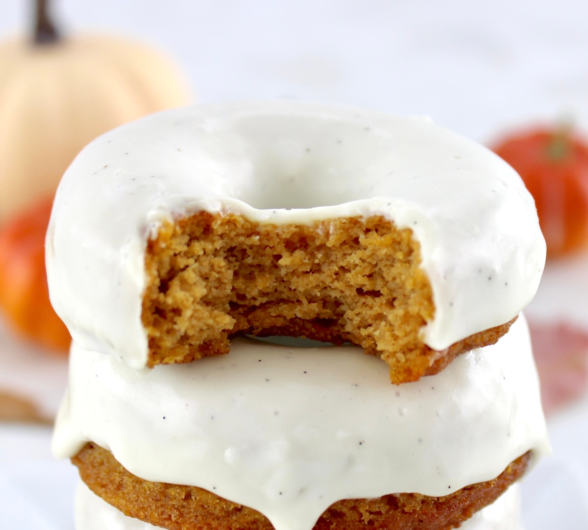 closeup of 2 Keto Pumpkin Donuts stacked with bite taken out of top donut with pumpkins in back
