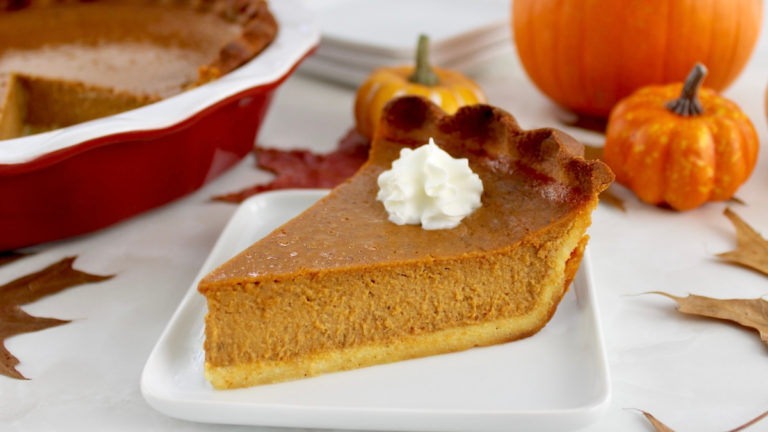 slice of Keto Pumpkin Pie on white plate with pumpkins in back