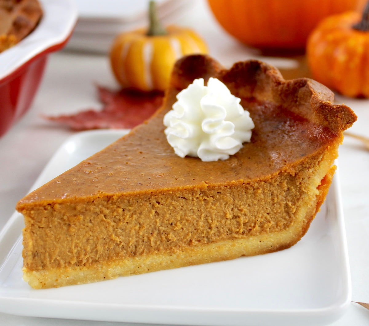slice of Keto Pumpkin Pie on white plate with pumpkins in back