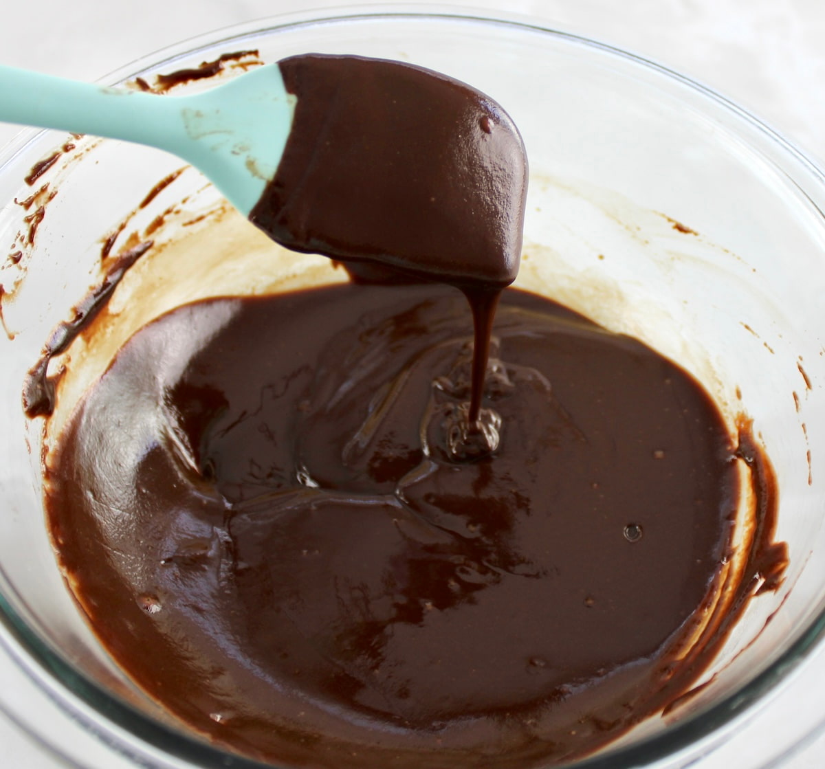 melted chocolate dripping off blue spatula in glass bowl