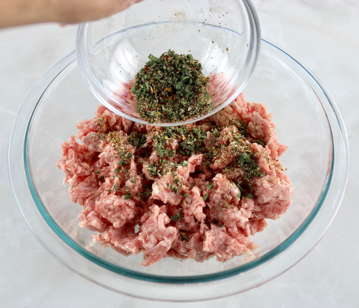 spices being poured over ground pork in glass bowl