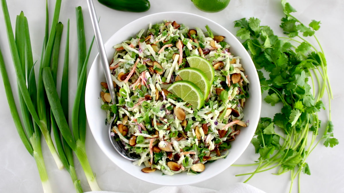 overhead view of Lime Cilantro Broccoli Slaw in white bowl with serving spoon and 3 lime slices on top