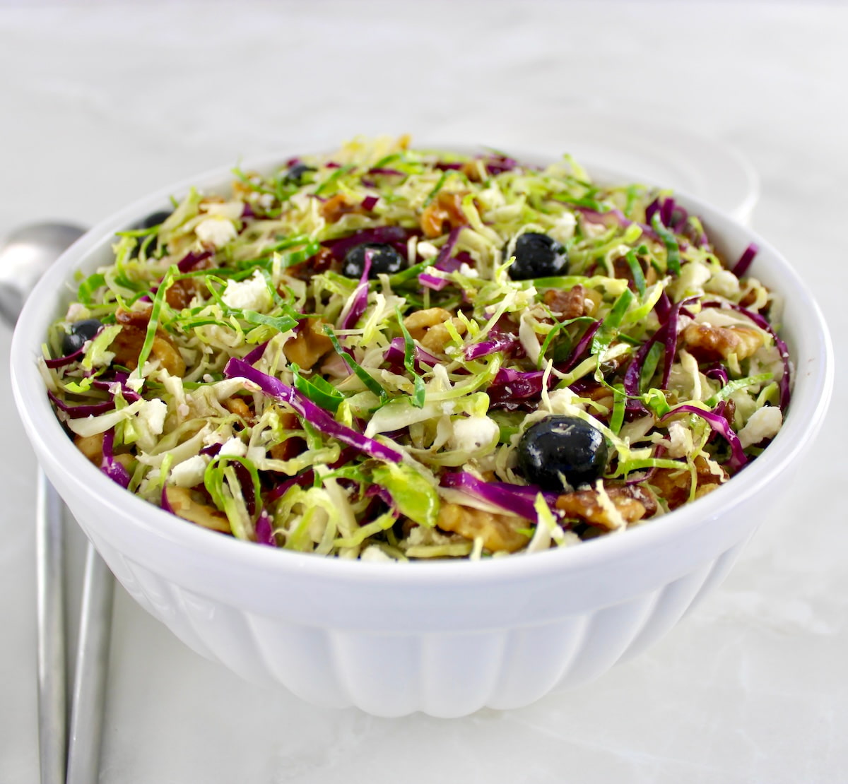 shaved brussels sprouts salad in white bowl with serving utensils on side