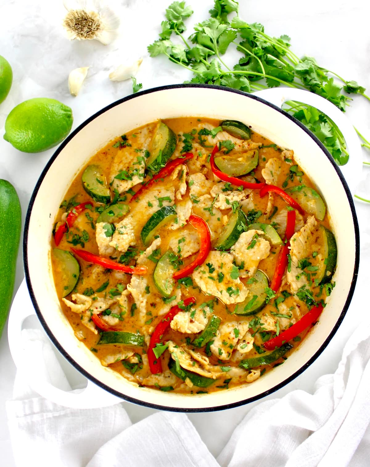 overhead view of Thai Red Curry Coconut Pork in white pan with cilantro, garlic and lime in back