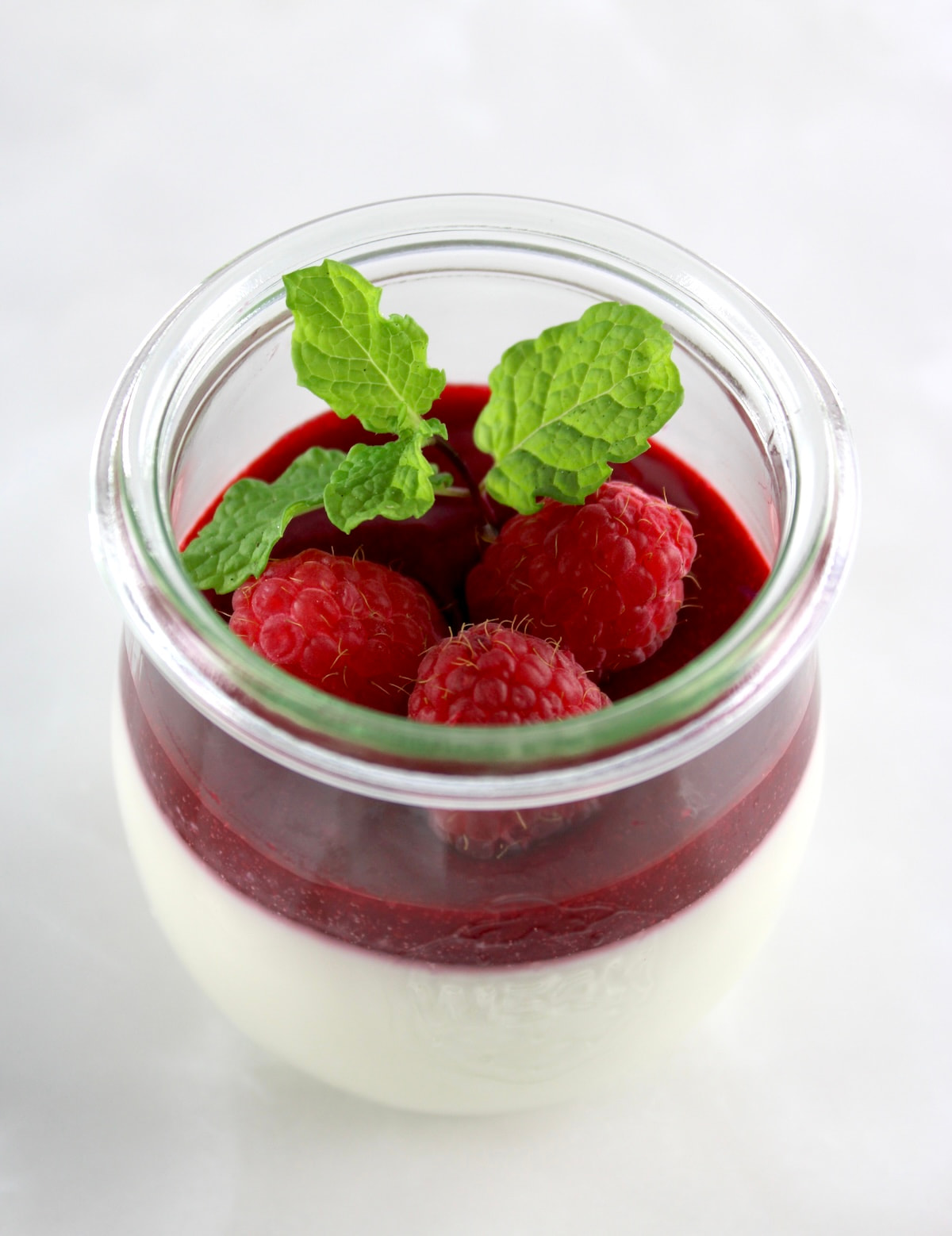 Keto Panna Cotta in open glass jar with raspberry sauce, fresh raspberries and mint on top