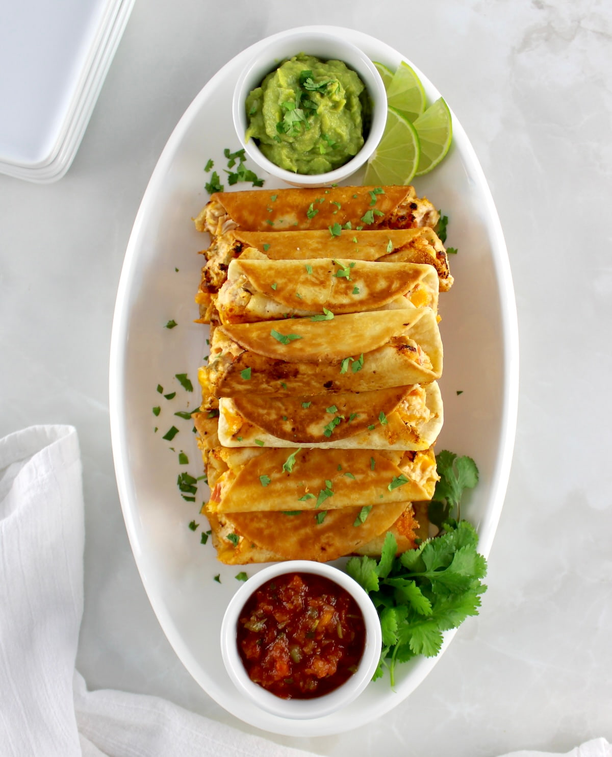 overhead view of Mini Chicken Taquitos on white oval platter with guacamole and salsa in white cups