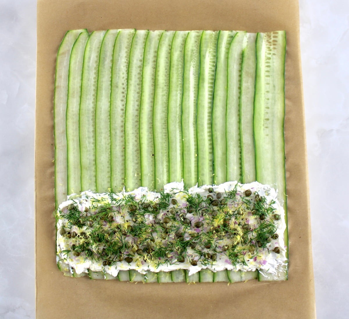 thin slices of cucumber on parchment with cream cheese, dill and capers on bottom end